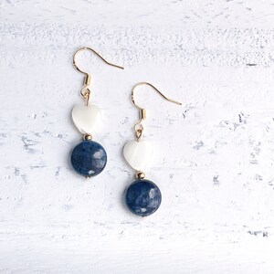 May include: A pair of dangle earrings featuring gold-colored hooks. Each earring has a white heart-shaped bead above a faceted, dark blue, round bead. The earrings are displayed on a white, textured surface.