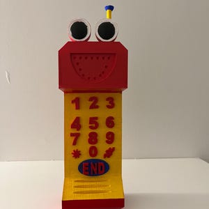 May include: A whimsical, toy phone in red, yellow, and blue. The top features large googly eyes and a small antenna. The phone has a keypad with numbers and the word "END".