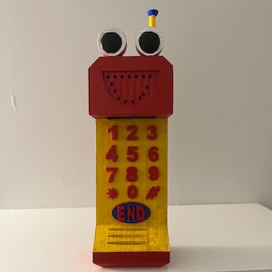 May include: A whimsical, handmade wooden toy phone with a red head featuring googly eyes and a smiling mouth. The yellow body has red number buttons and the word "END" in a blue oval.