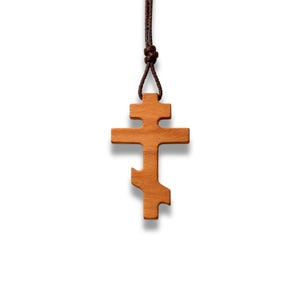 May include: A wooden Orthodox cross pendant with a brown cord. The cross has a three-bar design and is made of light brown wood. The cord is knotted at the top for wearing. The pendant is displayed against a white background.