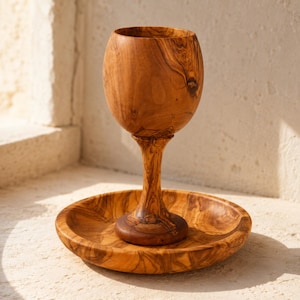 May include: A wooden goblet with a rounded bowl and a slender stem, resting on a matching circular wooden plate. The set features a distinct natural grain pattern in shades of light and dark brown, showcasing the organic texture of the wood.