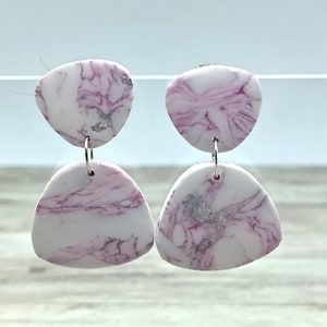 Puede incluir: Un par de pendientes colgantes con un diseño veteado en blanco y rosa. Los pendientes tienen un acento plateado y están conectados por aros plateados. Un accesorio único.