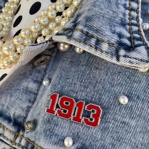 May include: A light-wash denim jacket adorned with faux pearls and a red and white enamel pin that reads "1913". The jacket is embellished with pearl-like beads and silver-toned buttons. A multi-strand pearl necklace is draped over a black and white polka dot surface.