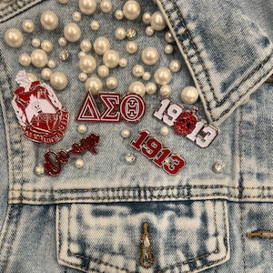 Op de afbeelding: Lichtblauwe spijkerjas versierd met imitatieparels en decoratieve spelden. De spelden hebben rood-witte emaille ontwerpen, Griekse letters en het jaar 1913. De jas heeft een klassiek ontwerp met knopen en voorzakken.