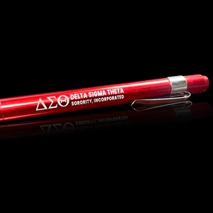 May include: A red pen with a silver clip. The pen has the text "DELTA SIGMA THETA SORORITY, INCORPORATED" printed in white. The pen is on a black surface.