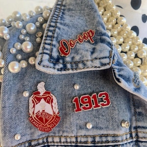 Op de afbeelding: Een lichtblauwe spijkerjas versierd met imitatieparels en spelden. De spelden bevatten de rode tekst "Do-oop", een wapen en het nummer "1913". De jas heeft een lichtblauwe wassing.