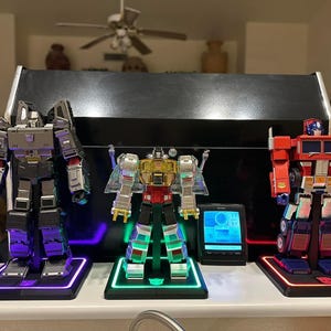 May include: Three Transformers action figures on display stands with LED lights. The figures are Megatron, Optimus Prime, and Bumblebee. Megatron is a dark gray and silver robot with a purple light. Optimus Prime is a red, yellow, and silver robot with a green light. Bumblebee is a red and silver robot with a red light.