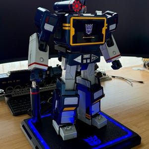 May include: A blue, grey, and white robot action figure standing on a black display base with blue LED lighting. The robot features a yellow-bordered chest screen with a purple Decepticon logo, a shoulder-mounted cannon, and playback control buttons.