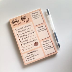 May include: A peach-colored notepad with the words "hello fall lets get it" written in brown script. The notepad has a list of tasks and goals for the fall season, including "schedule meetings", "finish editing drafts", "package stickies", "get groceries", "car wash", and "cancel free subscription". There are three goals listed, each with a number and a short description. The notepad also includes a section for "Top 3 Priorities" with three items listed.
