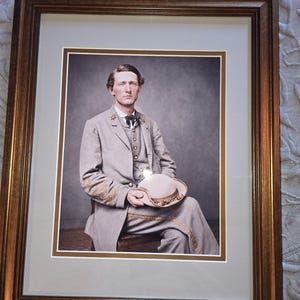 May include: A framed portrait of a person in a light gray military-style uniform with gold embroidery on the sleeves and collar, a vest with buttons, and a dark necktie. The person is seated and holds a light-colored hat in their lap.