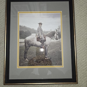 Puede incluir: Fotografía enmarcada en negro y dorado de un hombre a caballo. La imagen está en escala de grises, con un borde amarillo alrededor de la fotografía. El marco es negro con un borde interior dorado.