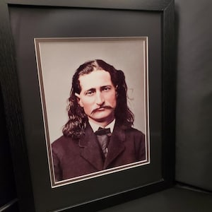 Puede incluir: Retrato enmarcado de un hombre con cabello largo y oscuro ondulado y un bigote prominente. Lleva un traje oscuro con pajarita. El retrato está en un marco negro con un passe-partout blanco.