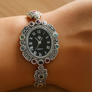 Puede incluir: Reloj de cuarzo plateado con esfera negra y números romanos. El reloj presenta un bisel decorativo con pequeñas piedras preciosas rojas y verdes. La correa del reloj también es plateada y está adornada con piedras preciosas similares.