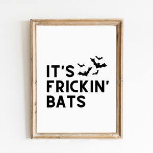 May include: A framed print with a white background and black text that reads "IT'S FRICKIN' BATS" with bat silhouettes. The frame is a light brown color and the print is displayed on a white wall.
