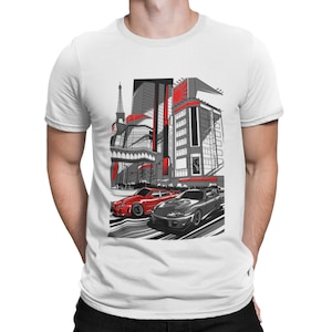 May include: White t-shirt with a graphic of two sports cars racing through a city street scene. The cars are red and gray, and the city buildings are in black and white. The graphic includes a red rising sun symbol.