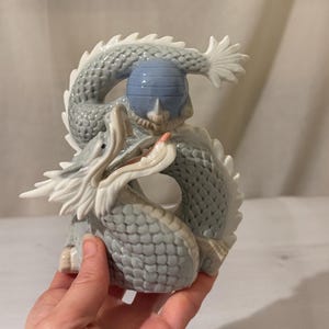 May include: A porcelain dragon figurine featuring a pale blue and white color palette with a textured scale pattern. The dragon is coiled in a circular shape, with its head turned upward, displaying white horns, a pink tongue, and a smooth blue sphere.