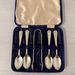 Pode incluir: Um conjunto de quatro colheres prateadas e uma pinça de açúcar a condizer, apresentadas num estojo azul com dobradiças e forro de cetim creme. Cada cabo de colher apresenta um emblema de coroa e gravações decorativas.