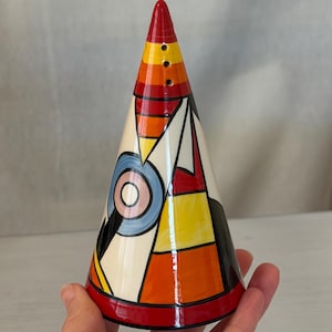 May include: A hand-painted ceramic salt shaker in a cone shape. The design features geometric shapes in red, orange, yellow, black, white, and blue. The top is red with three small holes for dispensing salt. The base has a red band.