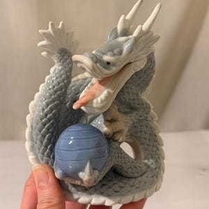 May include: A glazed porcelain figurine of a dragon in shades of light blue, gray, and white. The dragon has an open mouth with a visible pink tongue, white horns, and textured scales, while clutching a smooth, round, light blue sphere in its claws.