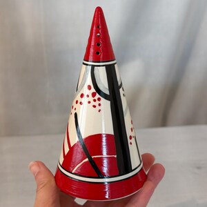 May include: A hand-painted ceramic cone-shaped object with a red top and base. The body is white with black and red abstract designs. The top has four small holes. The object is held in a hand.