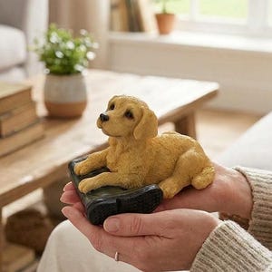 Puede incluir: Una figura de cachorro de color dorado que descansa sobre una bota de color verde oscuro. El cachorro tiene pelaje texturizado, nariz negra y mira hacia arriba. La figura se sostiene con dos manos, mostrando la forma detallada del perro.