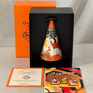 May include: A conical ceramic sugar sifter featuring a colourful landscape design with houses and trees, housed in a black display box with an orange outer sleeve. Includes a certificate of authenticity and a booklet titled Bizarre by Clarice Cliff.