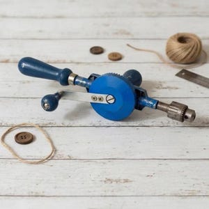May include: A vintage blue hand drill with a wooden handle and metal components. The drill is positioned on a white wooden surface, accompanied by buttons, twine, and a metal ruler. A classic tool for woodworking and crafts.