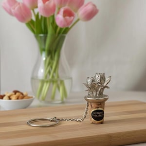 May include: A silver-toned bottle stopper with a floral design, attached to a chain and ring, rests on a wooden cutting board. A bottle of red wine, a glass of wine, and a vase of pink tulips are in the background.