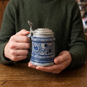 May include: A cylindrical stoneware beer stein featuring a blue and grey salt-glazed finish with intricate embossed floral scrollwork and geometric bands. The vessel is topped with a hinged pewter lid and includes a sturdy side handle.