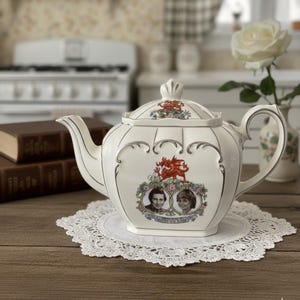 Peut inclure: Une théière en céramique blanche avec un couvercle, ornée de portraits et de motifs floraux. La théière a une bordure argentée et repose sur un napperon en dentelle. L'arrière-plan comprend une cuisine.