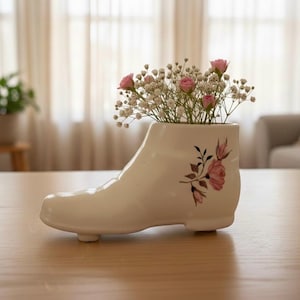 May include: A white ceramic boot-shaped vase filled with pink roses and white baby's breath. The vase has a floral design with pink roses and green leaves. The vase is on a wooden table.