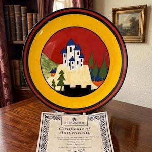May include: A decorative plate with a castle design. The plate features a red center with a yellow border and a black rim. The castle is white with blue roofs, and there are green trees. A certificate of authenticity is below the plate.