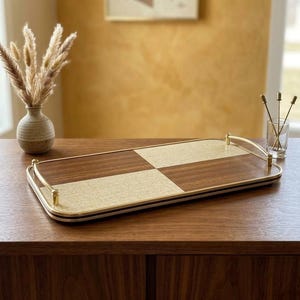 May include: A rectangular serving tray with a checkerboard design. The tray has a gold-coloured metal frame and handles, with alternating squares of dark wood and a light, textured material. The tray is on a wooden surface.