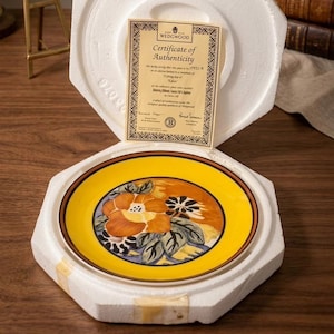 May include: A decorative plate with a floral design in shades of orange, yellow, and blue, set within a protective foam casing. The plate is marked with a brown border and a certificate of authenticity from Wedgwood.
