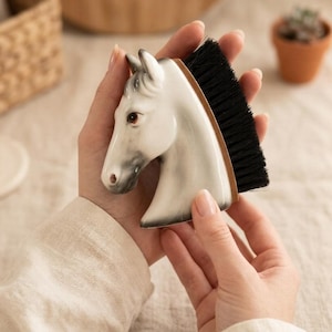 Puede incluir: Un cepillo con forma de cabeza de caballo de cerámica blanca con cerdas negras. La cabeza del caballo tiene sombreado gris y ojos marrones. El cepillo se sostiene con dos manos.