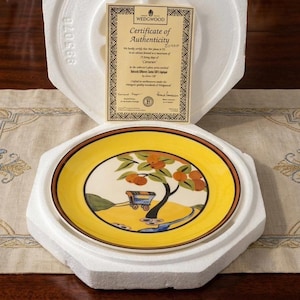 May include: A decorative plate with a yellow rim and a central design featuring a tree with orange-colored fruit and a small cart. The plate is in a protective foam casing, with a certificate of authenticity from Wedgwood.
