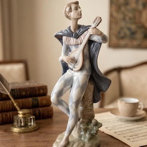 May include: A porcelain figurine of a musician in a harlequin costume, playing a lute. The figure wears a blue cape and a diamond-patterned outfit in white and light blue. The figurine is approximately 25 cm tall.