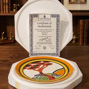 May include: A decorative plate from the Wedgwood collection, featuring a colorful tree design with orange, red, and blue accents. The plate is presented in a protective white foam case, accompanied by a certificate of authenticity.