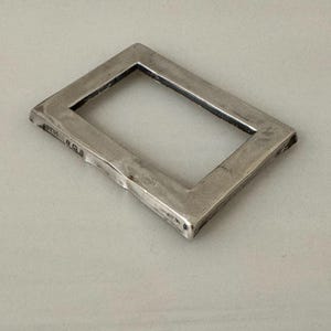 May include: A rectangular, silver-coloured metal frame with a hollow centre. The frame has a slightly textured surface and appears to be handmade. The corners are squared, and the edges are beveled. The frame is likely a decorative or functional item.