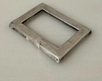 May include: A rectangular, silver-coloured metal frame with a hollow centre. The frame has a slightly textured surface and appears to be handmade. The corners are squared, and the edges are beveled. The frame is likely a decorative or functional item.