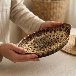 Könnte beinhalten: Eine ovale Servierplatte aus Keramik mit gesprenkelter brauner und beigefarbener Glasur. Die Platte hat einen strukturierten Rand. Eine kleine Olivenölflasche befindet sich im Hintergrund. Die Platte wird von einer Person gehalten.