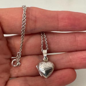 Puede incluir: Colgante de plata en forma de corazón con una delicada cadena. El medallón tiene un diseño grabado en la parte delantera. La cadena mide aproximadamente 45 cm de largo. El colgante está sujeto por una mano.