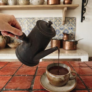 May include: A dark matte metal coffee pot pouring a stream of dark liquid into a speckled ceramic cup and saucer. The pot has a cylindrical body, a lid with a small knob, and a side handle. The cup is filled with a dark brown beverage.
