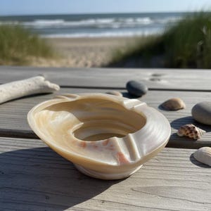 Puede incluir: Un cenicero beige en forma de concha con una hendidura circular y un borde curvo. El cenicero tiene un acabado brillante y está colocado sobre una superficie de madera, con una escena de playa al fondo. Conchas y guijarros están esparcidos alrededor del cenicero.