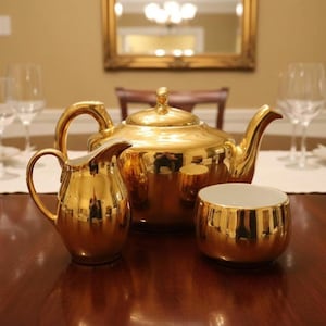 May include: A gold-colored tea set, including a teapot, creamer, and sugar bowl, is displayed on a dark wooden table. The set reflects the surrounding room, with a mirror and chandelier visible. Wine glasses and plates are also on the table.