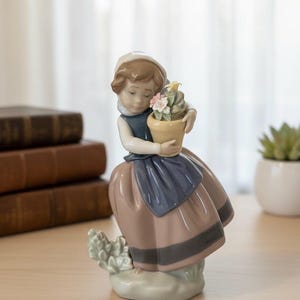 Può includere: Statuina in porcellana di una ragazza che tiene un vaso giallo con fiori rosa e bianchi. La ragazza indossa un vestito blu e marrone con un cappello bianco. La statuina è su una base verde chiaro.