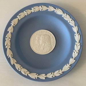 Puede incluir: Un plato decorativo azul con un borde de hojas de laurel blancas. El centro presenta un medallón plateado con los perfiles de dos hombres y el texto "THOMAS COKE - JOHN WESLEY".