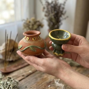 May include: A small terracotta pot with a textured rim, yellow circular accents, and a green vine relief, alongside a dark green glazed ceramic bowl featuring a gold-painted rim and base with three small drainage holes at the bottom.