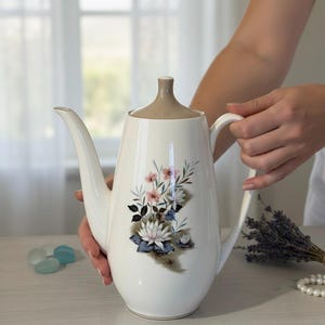 Peut inclure: Une cafetière en céramique blanche avec un couvercle et une poignée beiges. La cafetière est décorée d'un motif floral avec des fleurs roses, bleues et blanches. La cafetière est tenue par une personne.
