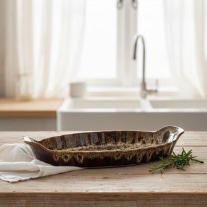 May include: A brown ceramic oval baking dish with a textured, speckled interior and handles. The dish is on a wooden table with a white cloth and sprig of rosemary. Kitchenware.